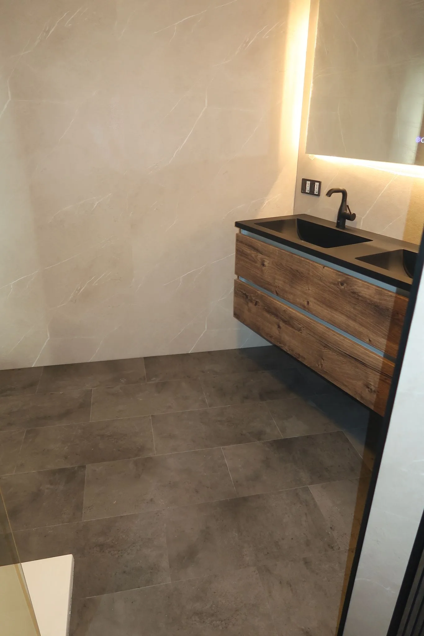 Een moderne badkamer met een houten wastafel, zwarte wasbak en donkere vloer. De muren zijn betegeld met grote witte tegels met marmerpatroon.